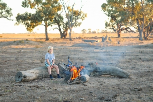 Farm boy sitting on log seat near camp fire in the Australian bush - Australian Stock Image