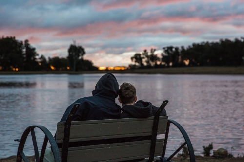 Father and son sitting on chair looking out over river water at sunset - Australian Stock Image