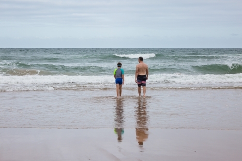 Father and son walking out towards waves at beach from behind - Australian Stock Image