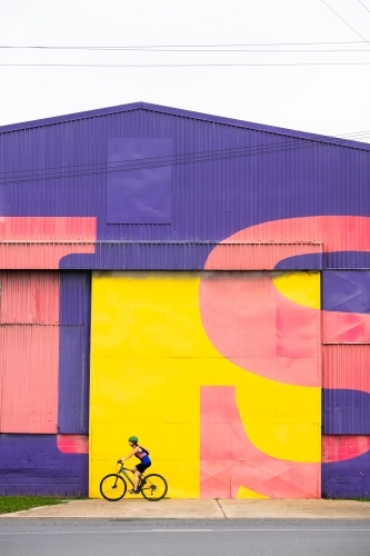 Female Cyclist Passing Bright Warehouse Wall - Australian Stock Image