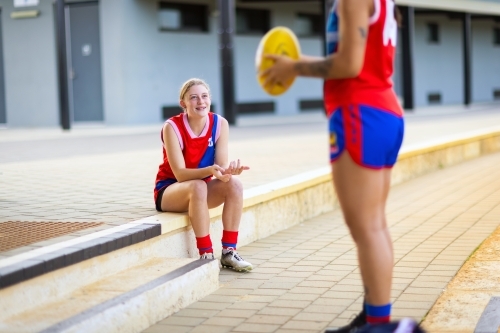 female football player sitting on step looking up at team member holding football - Australian Stock Image