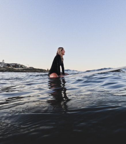 Female surfer sitting on longboard surfboard in springsuit with glassy blue ocean and clear sky - Australian Stock Image