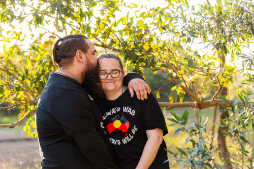 First Nations Australian couple embrace and husband kissing wife in Aussie bushland at sunset - Australian Stock Image