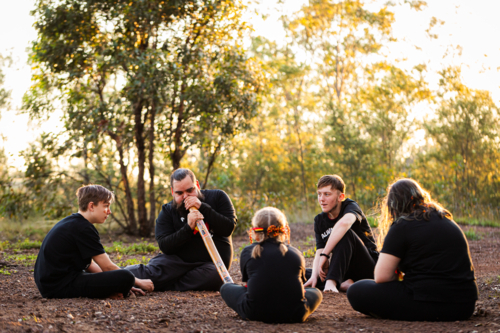 First Nations Australian man with his kids sitting in bushland playing didgeridoo - Australian Stock Image