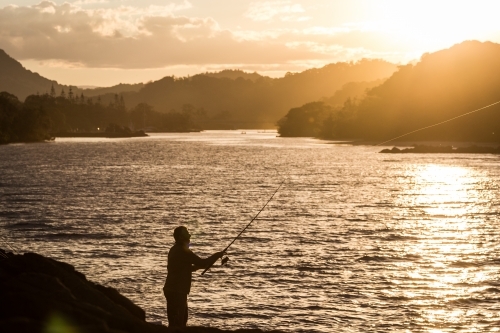 Fisherman standing on rocks beside a river at sunset - Australian Stock Image