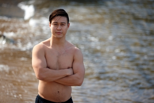 Fit Asian man folding arms at beach wearing only shorts - Australian Stock Image