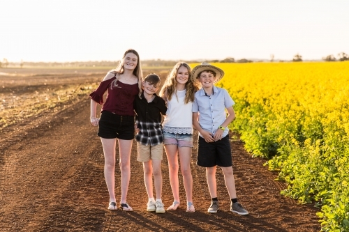 Four children standing close together happy smiling on farm near canola field - Australian Stock Image