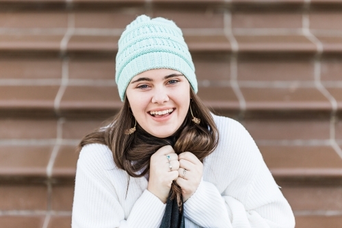 Girl clutching scarf around neck wearing beanie sitting on steps laughing - Australian Stock Image