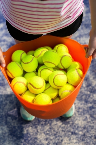 Girl holding an orange basket of tennis balls on a blue tennis court - Australian Stock Image