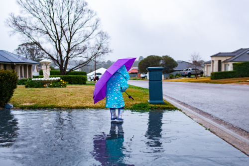 girl on rainy day wearing raincoat and holding purple umbrella splashing in puddle - Australian Stock Image