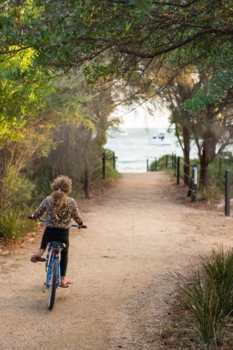 Girl riding a bike on gravel surrounded by trees and beach - Australian Stock Image