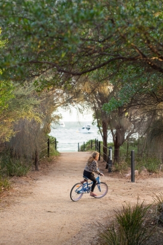 Girl riding a bike on gravel surrounded by trees and beach - Australian Stock Image