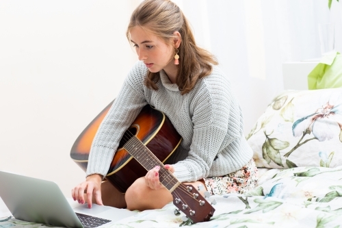 Girl sitting on bed with guitar looking at music on laptop - Australian Stock Image