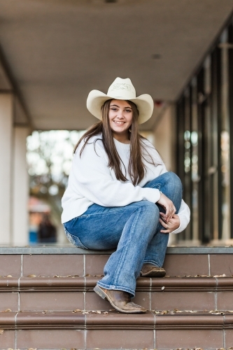 Girl sitting on steps with knees up wearing hat smiling - Australian Stock Image