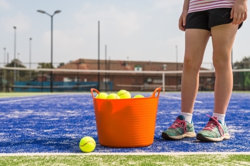 Girl standing next to an orange basket of tennis balls on a blue tennis court - Australian Stock Image