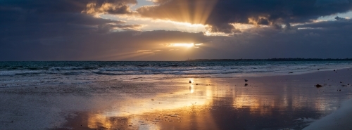 Golden sunshine breaking through dark clouds at sunset over a beach - Australian Stock Image