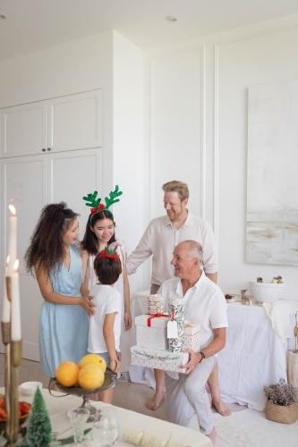 Grandfather sitting down holding Christmas presents while family smile and talk beside him - Australian Stock Image