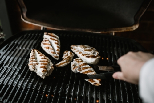 Grilled chicken breast on a BBQ being turned by a hand holding tongs - Australian Stock Image
