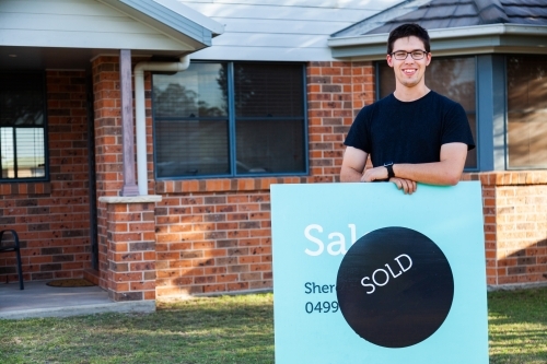 grinning young man with sold sign in front of newly purchased house - Australian Stock Image