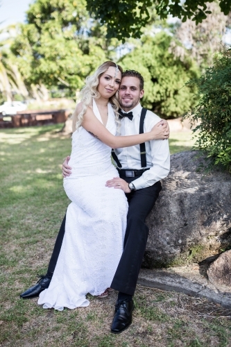 Groom sitting on rock with bride on his lap in garden - Australian Stock Image