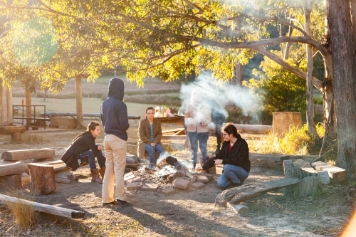 Group of young people relaxing around campfire in bushland on sunny winter morning