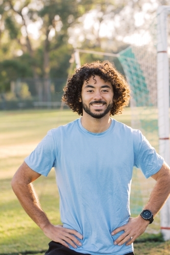 half body shot of a man with curly hair smiling with arms on his hips standing on a soccer field - Australian Stock Image