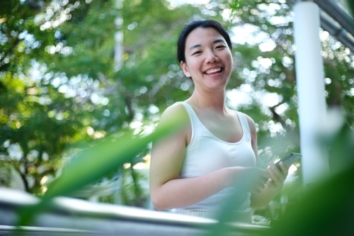 Happy Asian woman standing along walkway outdoors with green bokeh backdrop - Australian Stock Image