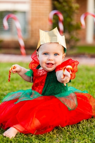 Happy baby at Christmas time sitting on lawn outside with candy canes and golden paper crown hat - Australian Stock Image