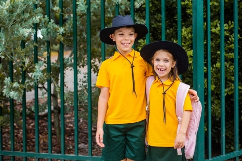 Happy brother and sister laugh and hug beside school fence outside - Australian Stock Image