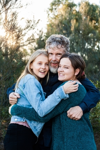 Happy dad with his two teenage daughters hug together outside - Australian Stock Image