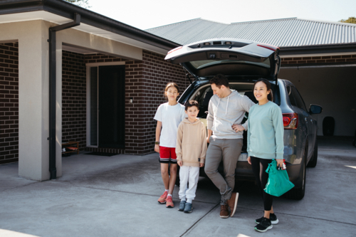 Happy family standing behind the car parked in the driveway with car boot open. - Australian Stock Image