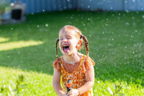 Happy laughing little girl joyfully playing with sprinkler hose watering herself in garden - Australian Stock Image