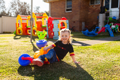 Happy little Aussie kid playing with colourful trike in backyard of home - Australian Stock Image