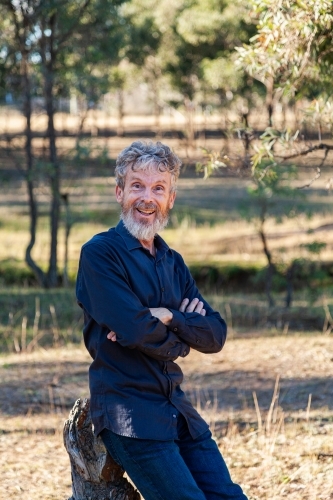 Happy middle aged man with beard leaning on stump in paddock - Australian Stock Image