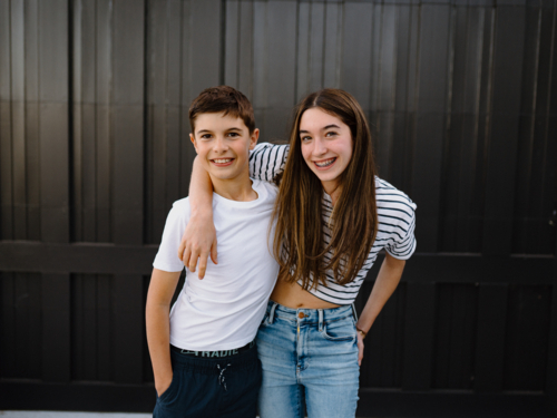 happy teen siblings standing outside against black garage door background - Australian Stock Image