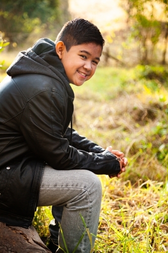 Happy tween boy sitting on log outside smiling - Australian Stock Image
