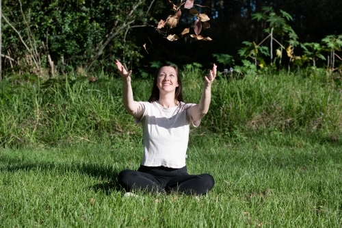 happy woman sitting cross legged in the grass tossing leaves in the air - Australian Stock Image