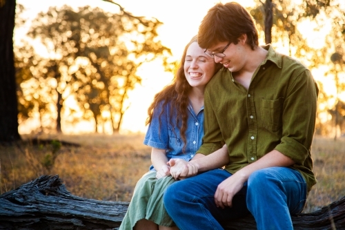 Happy young couple sitting on log at sunset smiling together - Australian Stock Image