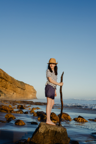 Happy young girl wearing a sunhat standing on a rock holding a stick playing at the beach - Australian Stock Image