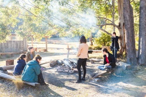 Happy young group of friends sitting around a campfire on a campground in the morning - Australian Stock Image