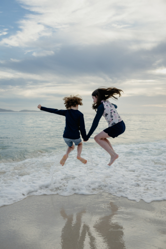 Happy young sister & brother (young girl and boy) holding hands jumping in the surf at the beach - Australian Stock Image