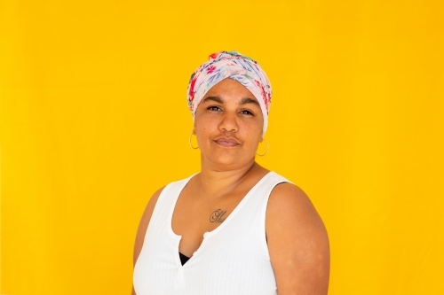 Head and shoulders of Indigenous woman wearing floral head wrap against bright yellow background - Australian Stock Image