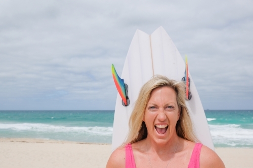 Head shot of Woman in bathers looking at camera on beach holding surfboard behind head - Australian Stock Image