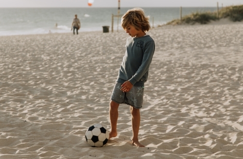 Horizontal shot of a boy playing with a soccer ball in the sand on the beach - Australian Stock Image