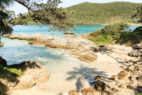 Horizontal shot of a remote beach with rocks mountains and trees on a sunny day with clear skies - Australian Stock Image