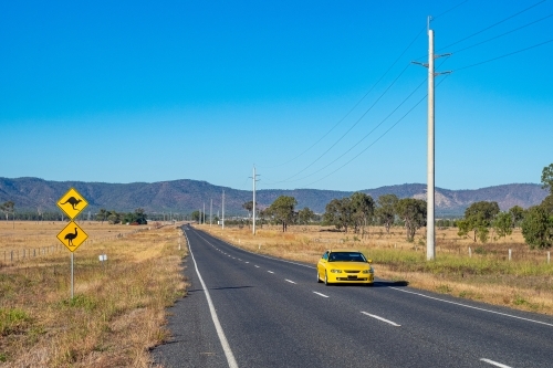 Horizontal shot of a yellow kangaroo and emu signage on the roadside and a highway with a yellow car - Australian Stock Image