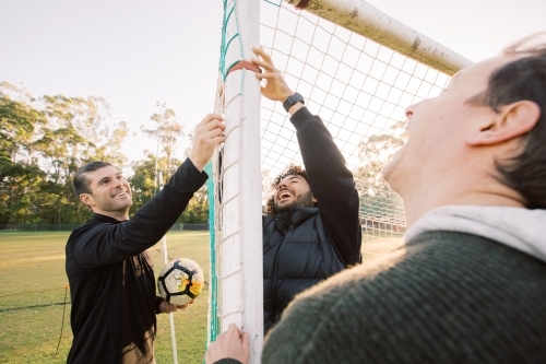 horizontal shot of three men smiling one holding a soccer ball and two touching the net - Australian Stock Image