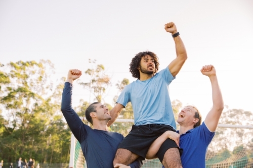 horizontal shot of two men carrying a man on their shoulders while raising their arms up high - Australian Stock Image