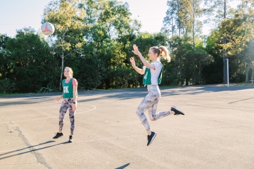 horizontal shot of two young women playing net ball with one jumping in mid air on a sunny day - Australian Stock Image