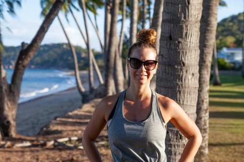 horizontal shot of woman smiling with sunglasses on by the beach with trees, mountains and waves - Australian Stock Image